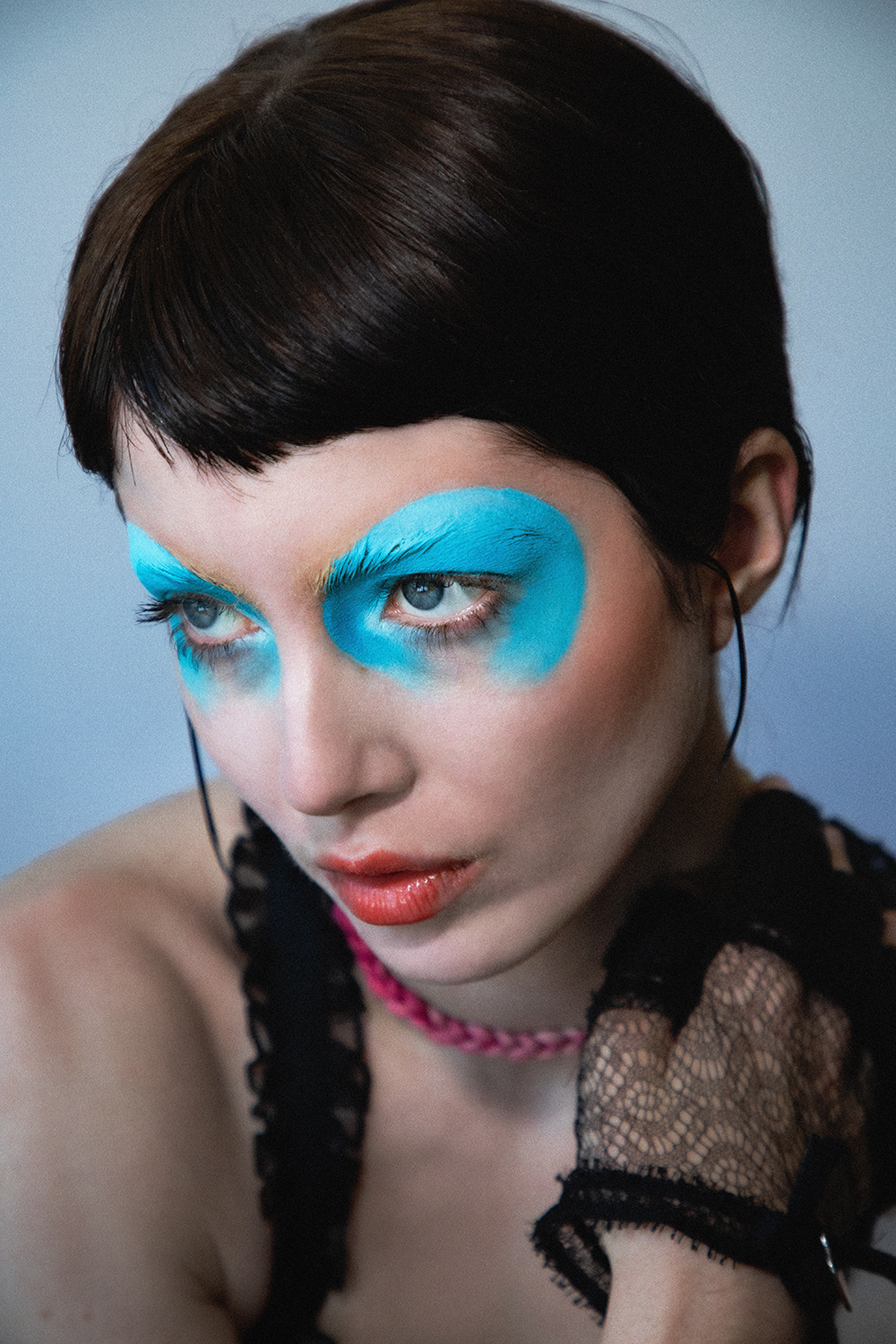 Close up portrait of Autry with bright blue graphic eye makeup looking past the camera with one hand on their shoulder